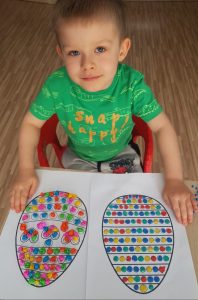 Kilkuletni chłopiec pokazuje dwie kartki z wyklejonymi pisankami. Jedna jest wyklejona kulkami plasteliny, druga wyklejona kulkami z bibuły.